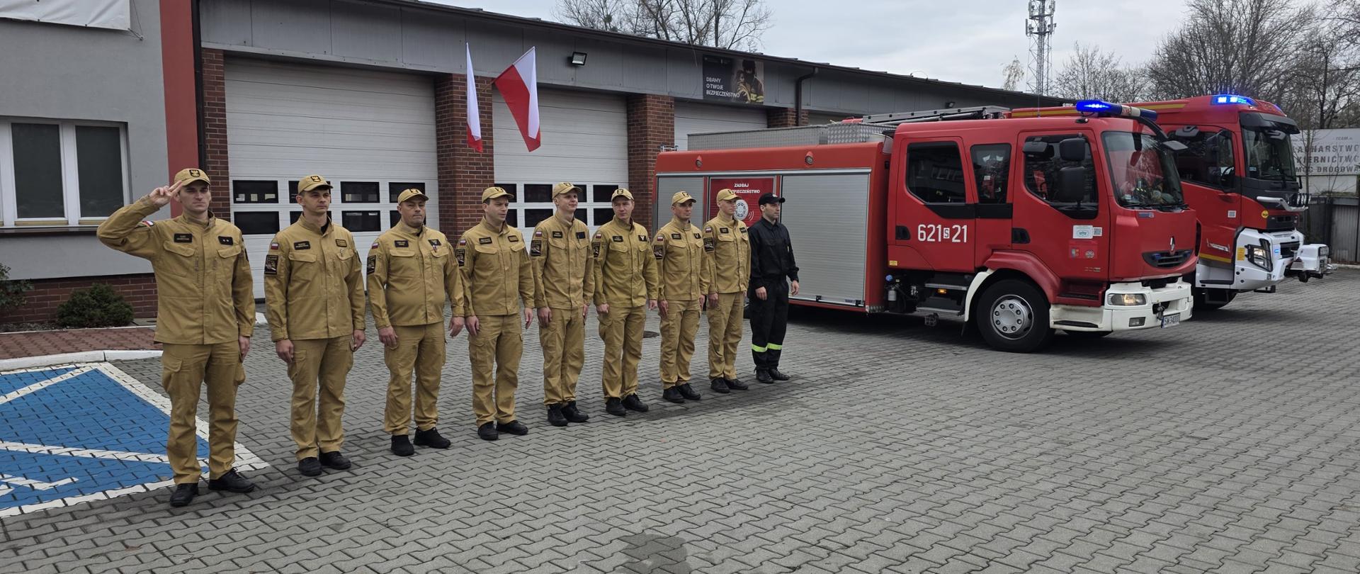Zdjęcie przedstawia uroczystą zbiórkę grupy strażaków w piaskowych mundurach bojowych, stojących w szeregu na placu przed remizą, z dwoma czerwonymi wozami strażackimi zaparkowanymi po prawej stronie. Scena ma miejsce w pochmurny dzień, a w tle widać bramy garażowe remizy oraz maszt z polską flagą.
