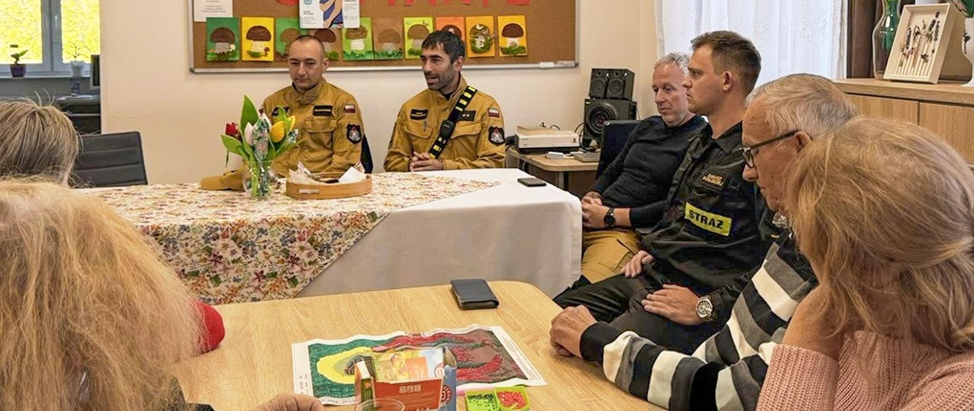 Zdjęcie przedstawia funkcjonariuszy PSP którzy siedzą przy stole. Przy drugim stole siedzą seniorzy.