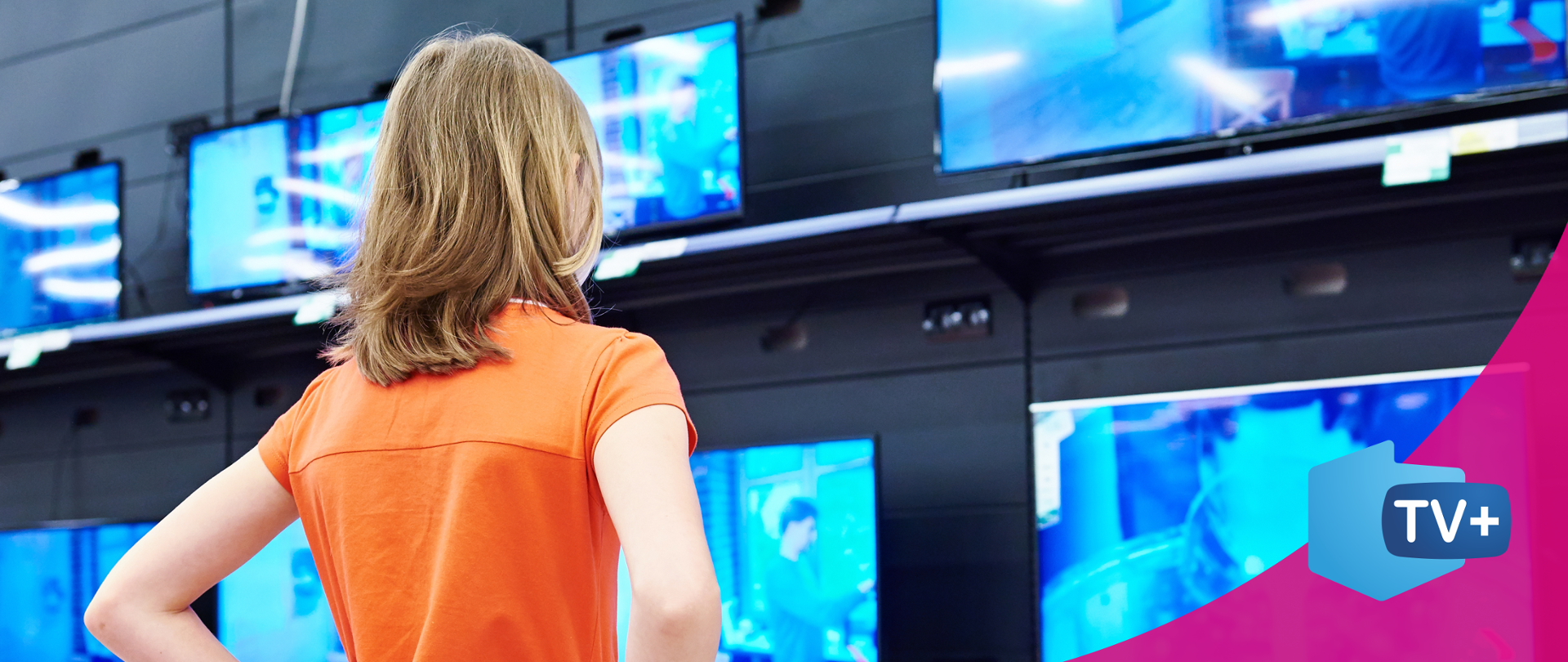 zdjęcie przedstawiające kobietę w pomarańczowej koszulce, stojącą tyłem. Patrzy w kierunku telewizorów wiszących na ścianie,.