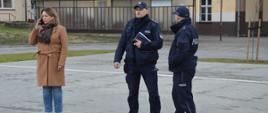 Ćwiczenia w ZSZ. Na parkingu szkolnym stoi dyrektor szkoły, która rozmawia przez telefon, a obok niej w pewnej odległości znajduje się dwóch policjantów. Jeden z nich trzyma w lewej ręce notatnik. W tle budynki szkoły. 