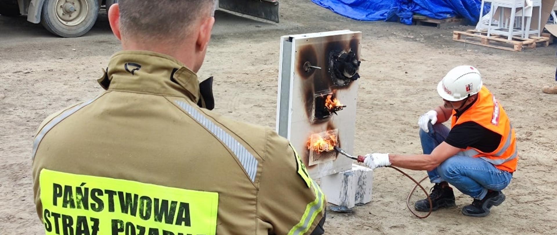 Na pierwszym planie zdjęcia widoczne plecy ubranego w piaskową kurtkę z napisem Państwowa Straż Pożarna mężczyzny bez nakrycia głowy. Przed nim schylony mężczyzna ubrany w jeansy i czarna koszulkę, w pomarańczowej kamizelce i białym kasku na głowie trzymający w lewej dłoni palnik. Obie dłonie w białych rękawicach ochronnych. Płomień z palnika obejmuje fragment stojącej przed nim ścianki z płyt. Niektóre elementy zwęglone od ognia, niektóre płoną. W tle piaszczyste podłoże placu, fragment naczepy, niebieska plandeka