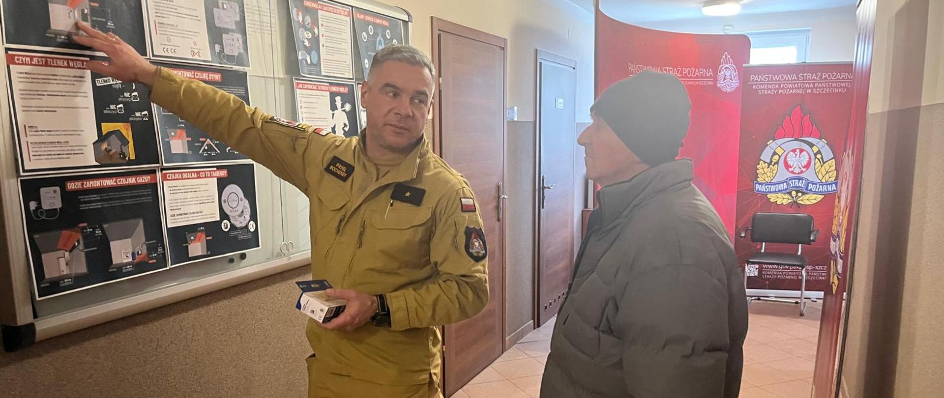 Strażak w żółtym mundurze w korytarzu budynku pokazuje na tablicy informacyjnej zasady dotyczące czujników tlenku węgla, trzymając w ręku niewielkie urządzenie, podczas gdy stojący obok mężczyzna w zimowej kurtce słucha wyjaśnień. W tle widoczny jest baner Państwowej Straży Pożarnej.