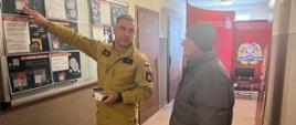Strażak w żółtym mundurze w korytarzu budynku pokazuje na tablicy informacyjnej zasady dotyczące czujników tlenku węgla, trzymając w ręku niewielkie urządzenie, podczas gdy stojący obok mężczyzna w zimowej kurtce słucha wyjaśnień. W tle widoczny jest baner Państwowej Straży Pożarnej.