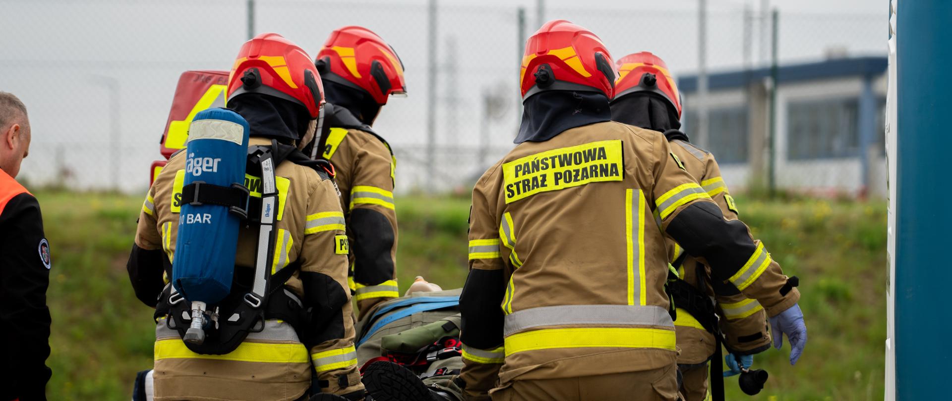 Strażacy: Na pierwszym planie widać czterech strażaków ubranych w pełne umundurowanie bojowe (piaskowe kurtki i spodnie z elementami odblaskowymi). Formacja należy do Państwowej Straży Pożarnej, co potwierdza wyraźny napis na plecach jednego z nich.
Działania ratownicze: Strażacy niosą na noszach osobę poszkodowaną (lub manekina szkoleniowego). Widoczne są jedynie nogi i fragment głowy osoby transportowanej.
Ekwipunek: * Jeden ze strażaków ma na plecach aparat oddechowy marki Dräger z butlą z napisem "300 BAR".
Wszyscy mają na głowach charakterystyczne czerwone hełmy strażackie.
W tle, na ziemi, widać rozłożoną srebrno-złotą folię termiczną (koc ratunkowy NRC).
Otoczenie: Akcja toczy się na zewnątrz, na utwardzonym podłożu (kostka brukowa) graniczącym z trawiastym nasypem. W tle widać ogrodzenie oraz fragmenty zabudowań technicznych, co sugeruje teren poligonu ćwiczebnego lub obiektu przemysłowego.