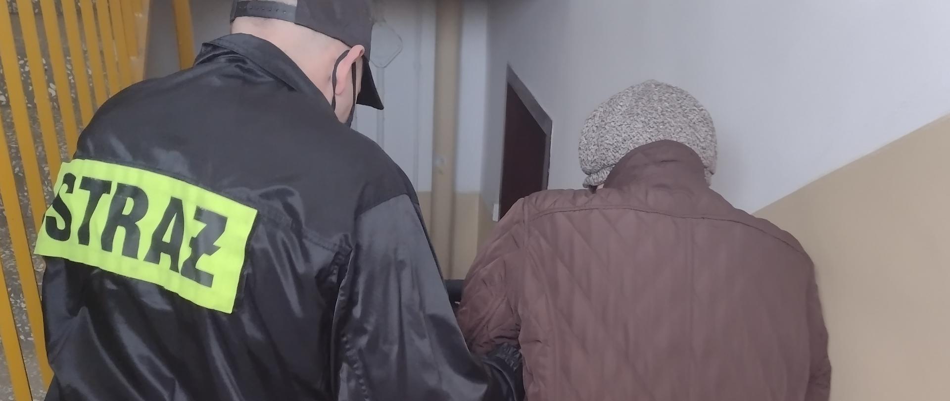 Na zdjęciu widać starszą kobietę oraz strażaka, który pomaga zejść kobiecie po schodach na klatce schodowej w budynku mieszkalnym wielorodzinnym
