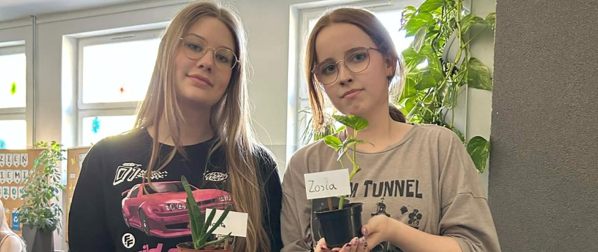 Dwie uczennice w okularach pozują na szkolnym korytarzu, trzymając w dłoniach małe doniczki z roślinami. Jedna roślina to aloes, druga to syngonium z karteczką z napisem "Zosia". W tle widoczna jest duża roślina pnąca na ścianie oraz gazeta ścienna z okazji Dnia Ziemi.