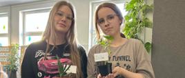 Dwie nastolatki w okularach pozują w jasnej sali szkolnej, trzymając w rękach małe doniczki z roślinami wyhodowanymi w ramach projektu szkolnego. W jednej z doniczek widoczna jest karteczka z imieniem rośliny. W tle widoczne zielone liście pnącza i gazetka ścienna o tematyce ekologicznej.