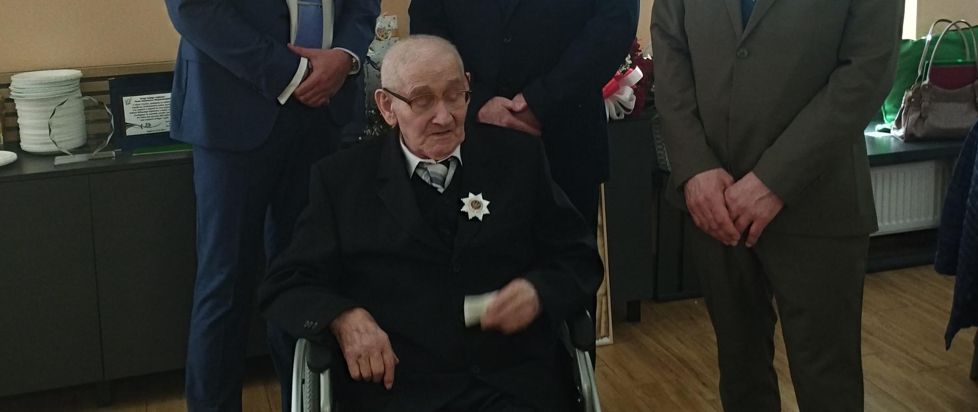 W sali pozują do zdjęcia trzej stojący mężczyźni w garniturach. Przed nimi na wózku inwalidzkim siedzi starszy mężczyzna z orderem w klapie marynarki. Jubilat jest przejęty podniosłą chwilą.