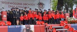 na scenie stoją dzieci z dziecięcej drużyny pożarniczej za nimi strażacy, a tyle sceny baner z napisem wojewódzkie obchody dnia strażaka