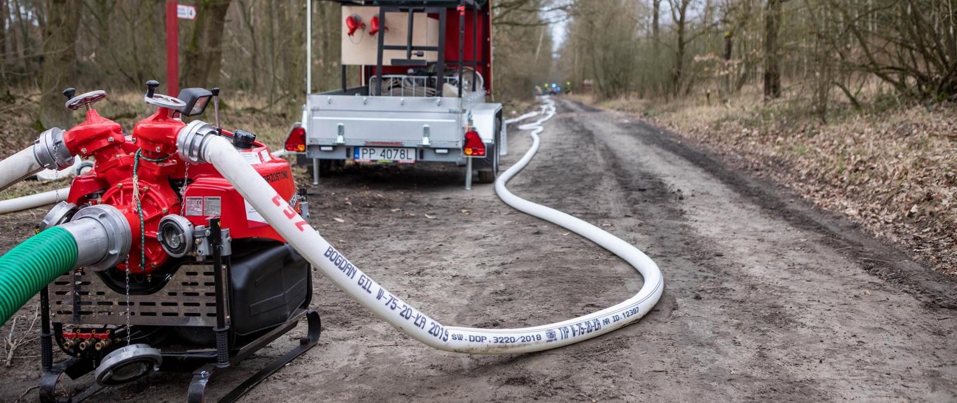 Na pierwszym planie zdjęcia stoi na leśnej drodze motopompa od której pociągnięte są linie wężowe.
