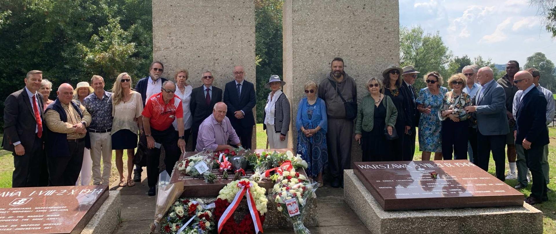 A group photo of 25 participants of the event posing in front of the Katyn Memorial; in the foreground three red graphite plaques; on the middle plaque lay several wreaths and bouquets of the white and red flowers 