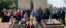 A group photo of 25 participants of the event posing in front of the Katyn Memorial; in the foreground three red graphite plaques; on the middle plaque lay several wreaths and bouquets of the white and red flowers 
