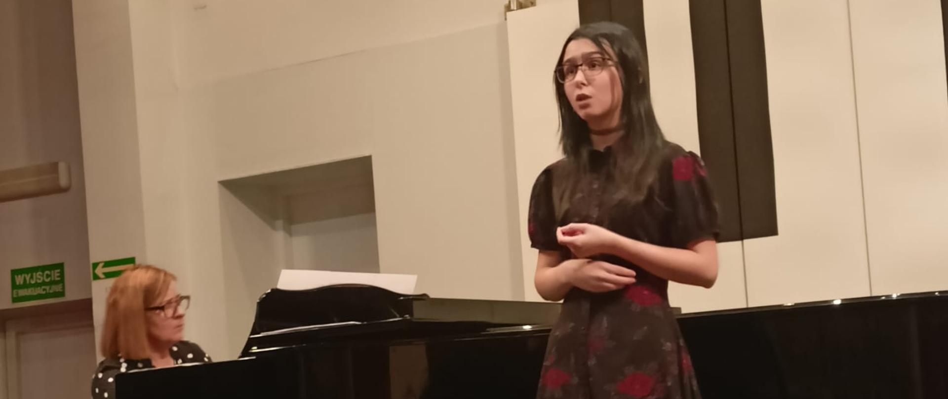 Dziewczyna śpiewa, kobieta akompaniuje jej na fortepianie.