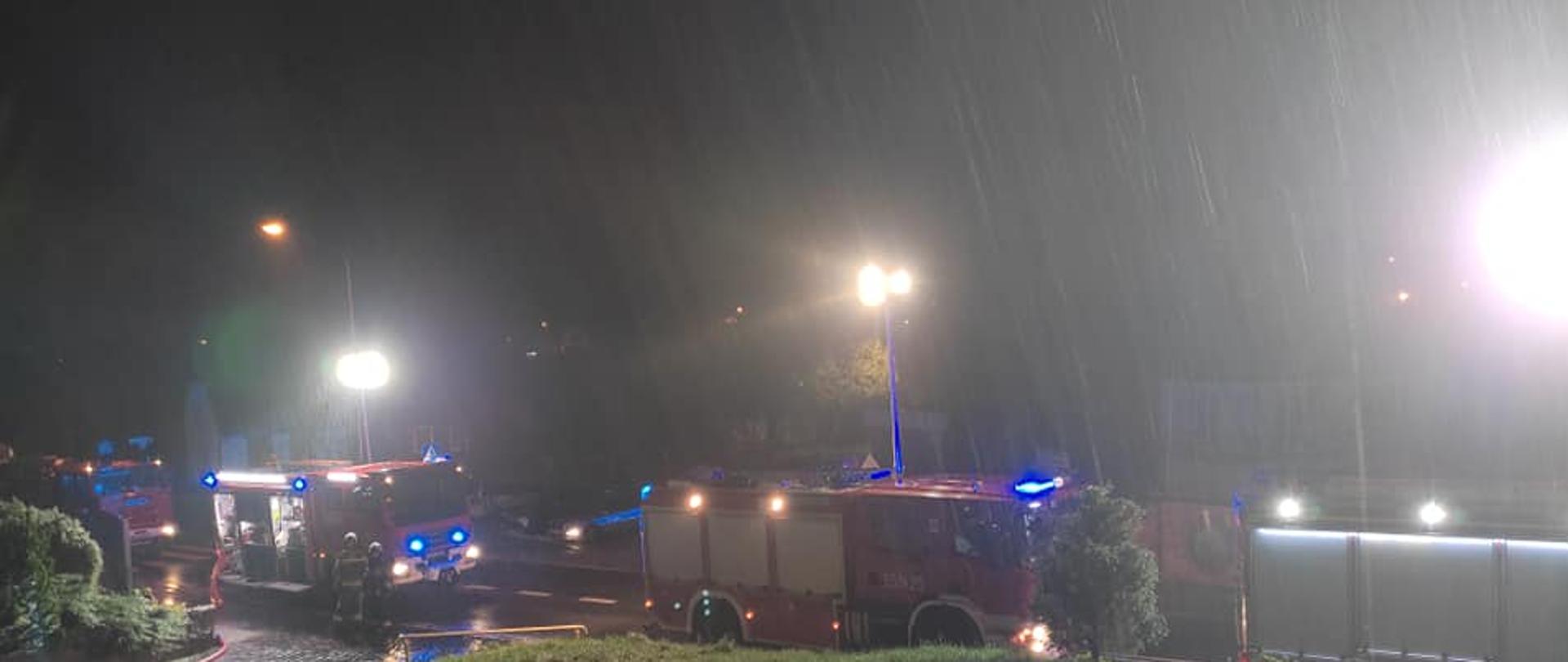 Zdjęcie przedstawia teren działań ratowniczych. Dwa wozy strażackie oświetlają teren akcji oraz podają wodę wężami.