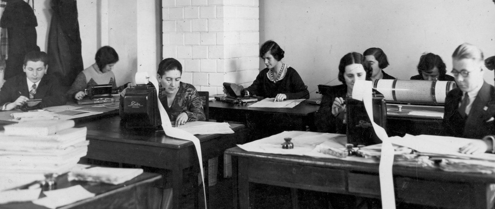 Na zdjęciu widzimy salę, w której znajdują się pracujący maszyniści i maszynistki, przy starych maszynach do pisania. Z tyłu sali jest wieszak z kurtkami oraz stary piec kaflowy. Na zdjęciu jest 6 pań maszynistek oraz 2 panów maszynistów. Na biurkach leżą papiery.