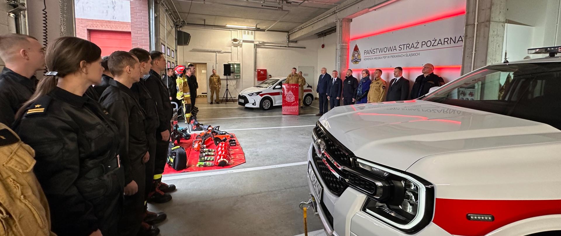 Na zdjęciu na pierwszym planie znajduje się przód białego samochodu ratowniczego Ford Ranger oraz strażacy komendy. W dalszym planie na czerwonej macie znajduje się sprzęt ratowniczy. W ostatnim planie widać zaproszonych gości, samochód operacyjny Toyota Yaris oraz mównicę z której przemawia komendant miejski. Wszyscy znajdują się w garażu jednostki ratowniczo - gaśniczej.