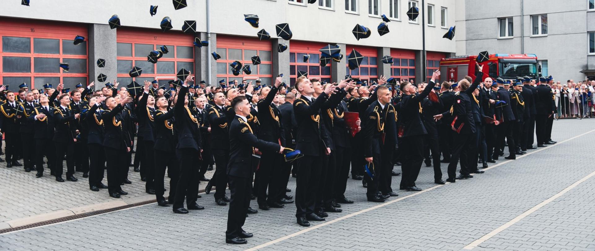 Nowo promowani aspiranci rzucają czapki w górę w geście radości na zakończenie ceremonii. W tle budynek szkoły z banerem "Promocja".