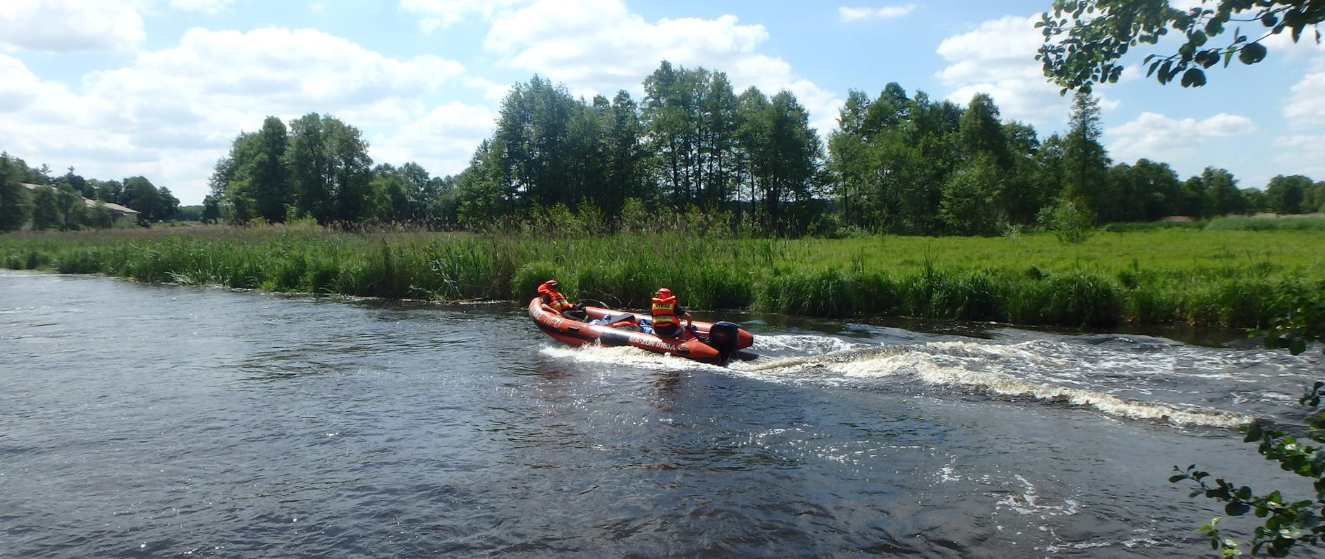 Łódką ratowniczą płyną strażacy na rzece. W tle drzewa.