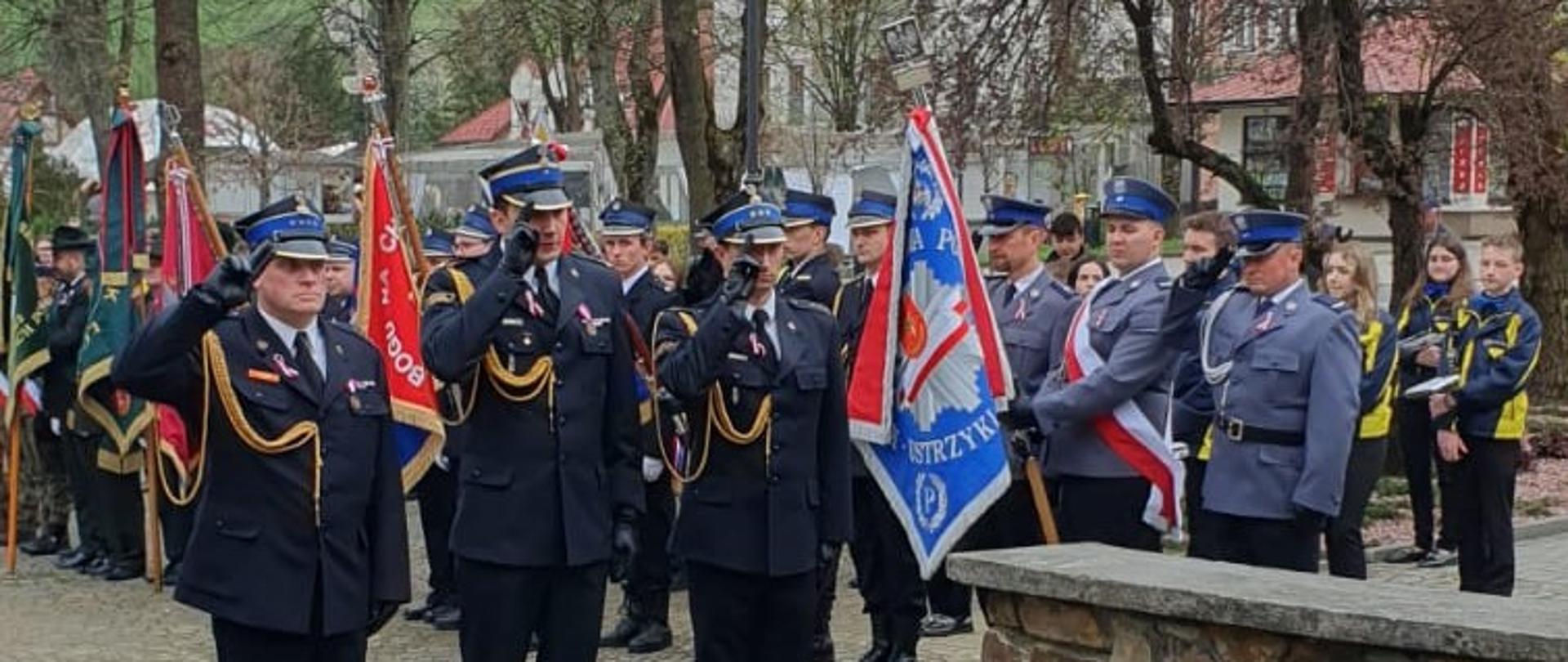 Na zdjęciu 3 oficerów z PSP w Ustrzykach Dolnych oddających honor przed pomnikiem na rynku w Ustrzykach Dolnych. W tle Poczty Sztandarowe.