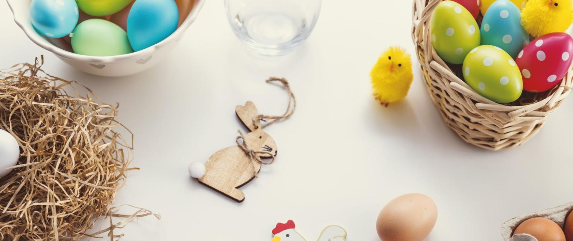 w centralnej części dłonie trzymają drewnianą kurkę, w górnej części jest drewniany królik oraz kolorowe pisanki w wiklinowym koszyku i porcelanowej misce