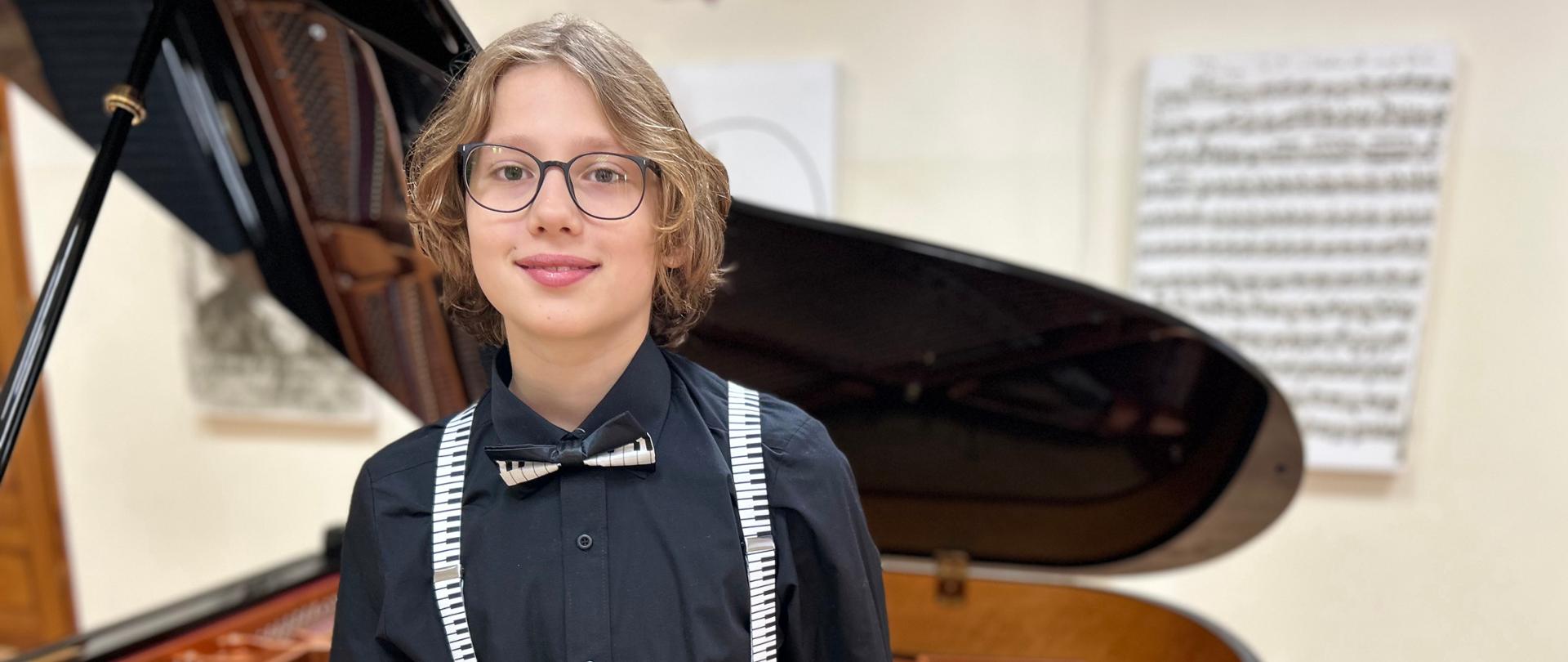 Zdjęcie przedstawia chłopca w okularach ubranego na czarno i opartego o czarny fortepian. W tle rozmazane grafiki o tematyce muzycznej. 