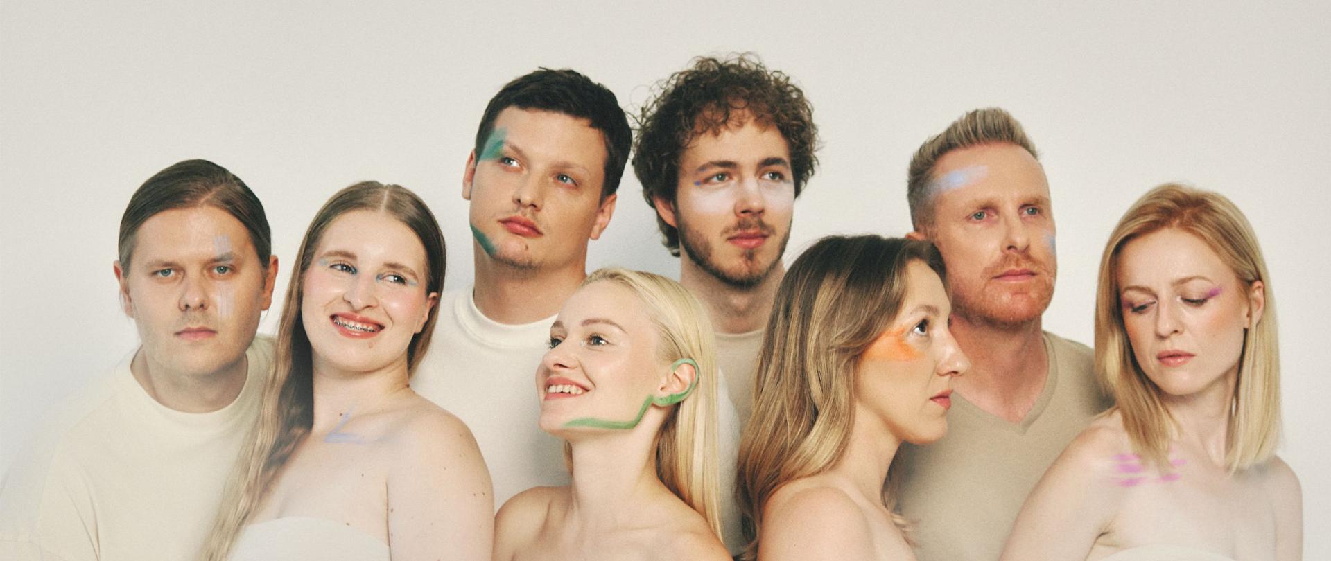 Grupo de oito pessoas alinhadas lado a lado sobre um fundo claro, retratadas do nível dos ombros para cima. Cada uma olha numa direção diferente e tem marcas suaves de tinta em tons pastel no rosto. As expressões variam entre sorriso e contemplação, criando uma composição artística e serena.