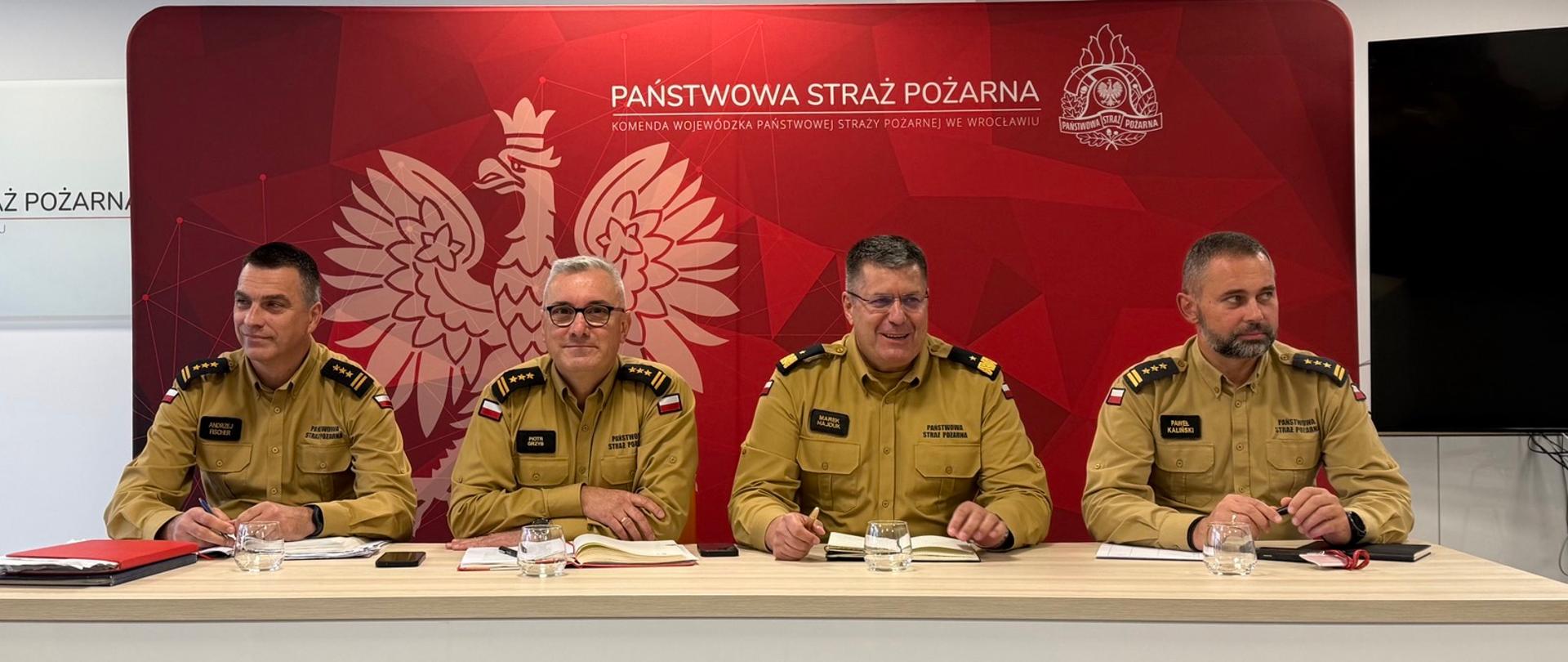 Czterech mężczyzn w galowych mundurach Państwowej Straży Pożarnej siedzi przy długim stole podczas konferencji prasowej. Za nimi znajduje się czerwona ścianka (tło) z napisem „PAŃSTWOWA STRAŻ POŻARNA” oraz godłem Polski i straży.