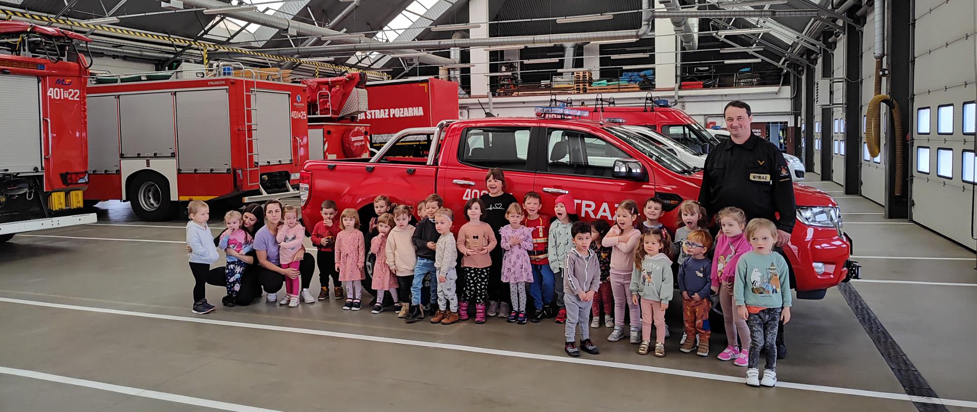 Zdjęcie grupowe dzieci i strażaka na tle samochodów pożarniczych w garażu JRG. 