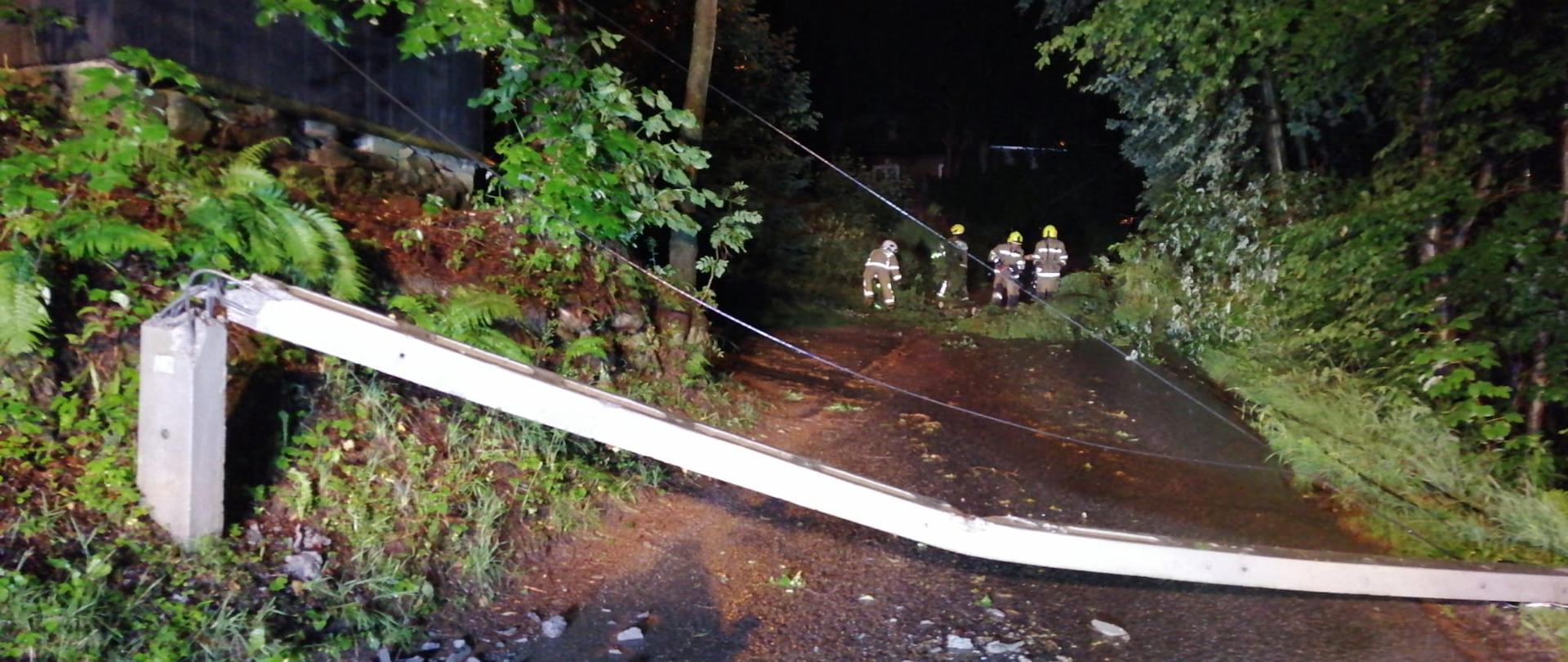 na zdjęciu strażacy usuwający w nocy złamane gałęzie z lokalnej drogi a na pierwszym planie przewrócony betonowy słup energetyczny