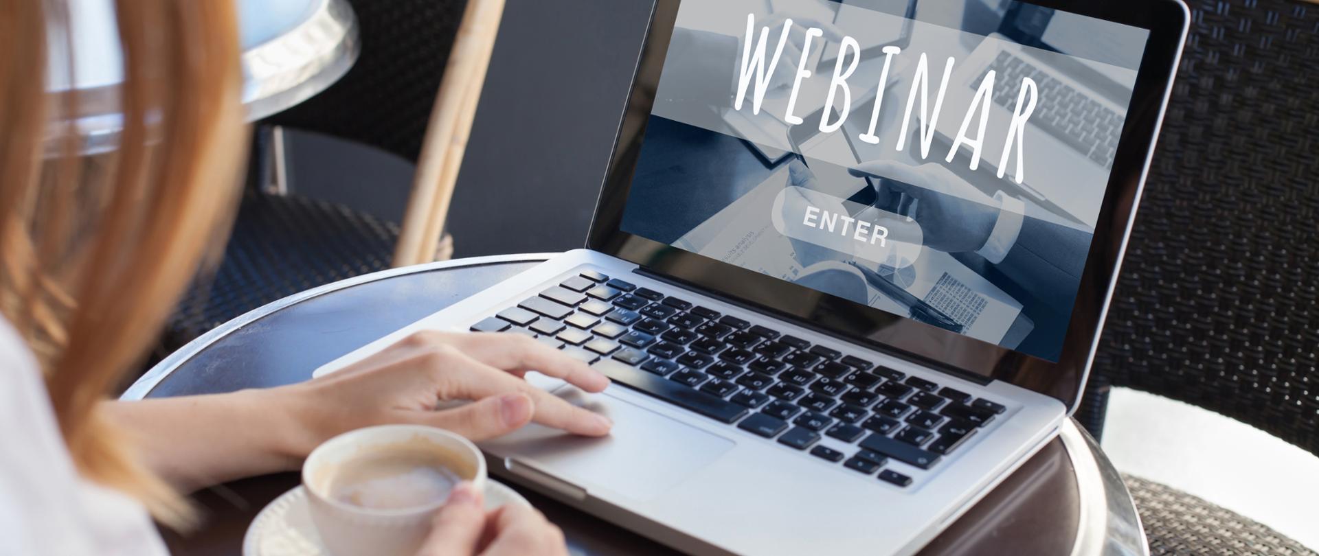 Kobieta siedząca przy stoliku z laptopem, w prawej ręce trzyma filiżankę z kawą. Na ekranie komputera napis webinar