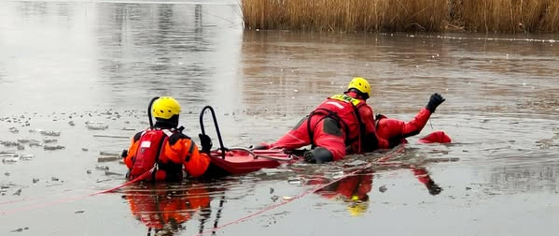 Strażacy w przeręblu zamarzniętego jeziora, trwają ćwiczenia, ubrani w specjalne kombinezony.