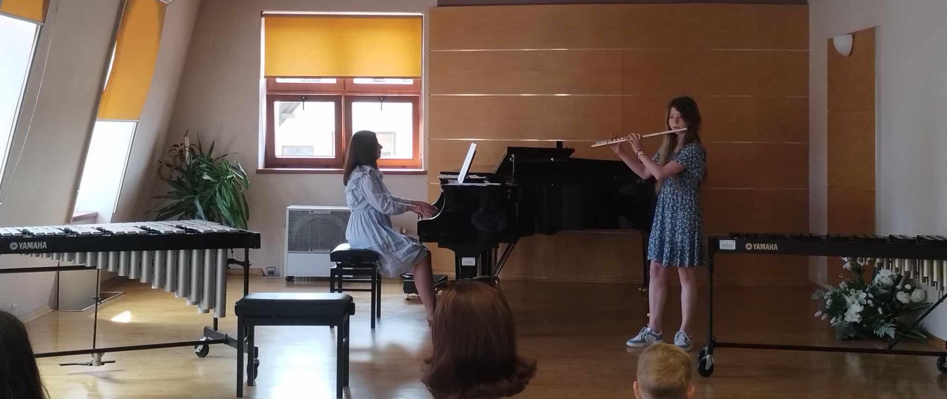 Na scenie sali kameralnej uczennica gra na flecie, obok niej akompaniatorka gra na fortepianie Z prawej i lewej strony sceny instrumenty perkusyjne. Za nimi trzy okna przysłonięte roletami pod jednym oknem nawilżacz powietrza. W rogu palma doniczkowa. Na pierwszym planie publiczność.