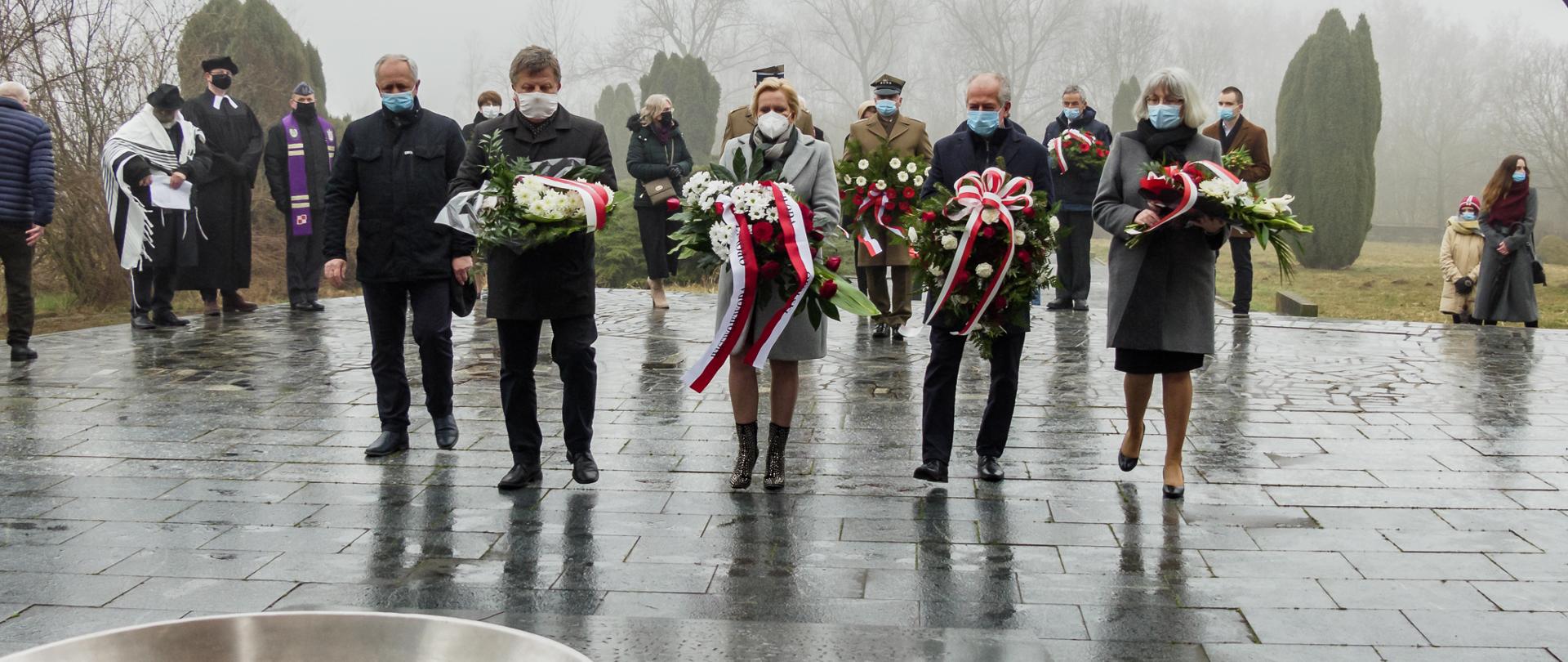 Na pierwszym planie 5 osób, idących kolumną na przeciw pomnika w Łambinowicach. W środku kadru wicewojewoda opolski, trzymająca wieniec. W tle pozostali uczestnicy uroczystości - widać m.in. żołnierzy. Na dole kadru w jego centralnej części widać tlący się płomień , wydobywający się z elementu ozdobnego pomnika. 