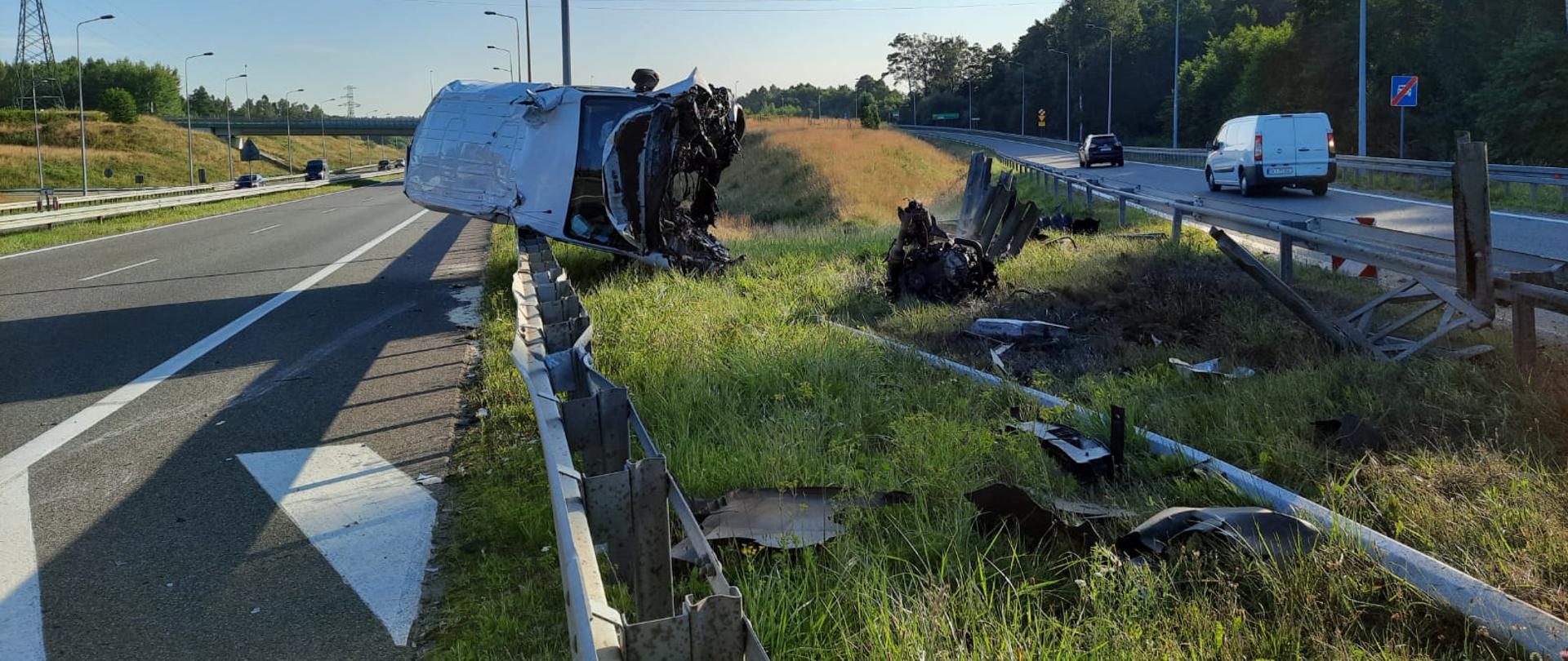 Bus leży na prawym boku między barierami energochłonnymi. Przed nim jest dużo elementów zgniecionej bariery. Za pojazdem ruch odbywa się płynnie na trasie S7. W Tle widać dalszą część trasy S7 i infrastrukturę drogową. Przed pojazdem leży silnik który na skutek bardzo silnego zderzenia wypadł z pojazdu.