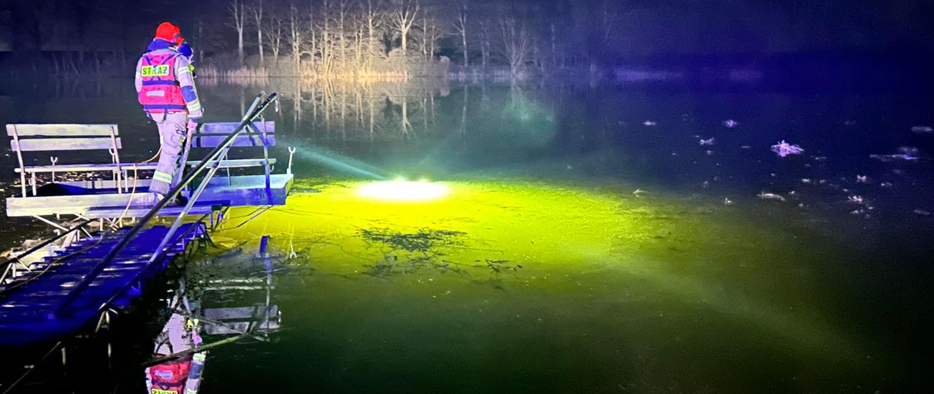 Noc, tafla lodu nad jeziorem. Strażak stoi na drewnianym pomoście.Spod lodu widać żółte światło drona podwodnego.