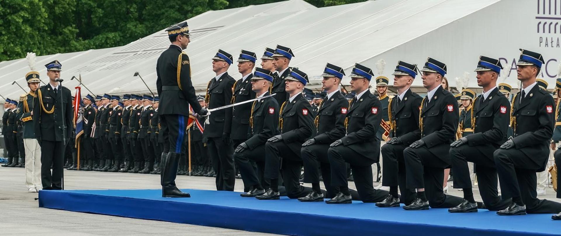 Zdjęcie przedstawia uroczystą ceremonię promowania oficerów Państwowej Straży Pożarnej, odbywającą się na świeżym powietrzu na placu apelowym. Na podwyższeniu przykrytym niebieskim dywanem stoi grupa strażaków w czarnych mundurach wyjściowych. Siedmiu strażaków klęczy na jedno kolano w równym rzędzie, z dłońmi spoczywającymi na kolanach. Mają na sobie czapki rogatywki z otokami. Przed nimi stoi wyższy rangą oficer, zwrócony do nich bokiem. Wykonuje on tradycyjny gest pasowania szablą, dotykając ostrzem ramienia jednego z klęczących strażaków. Oficer ma na sobie mundur z ozdobnymi sznurami oraz spodnie z niebieskimi lampasami wpuszczone w wysokie buty z cholewami. Za klęczącymi strażakami stoi rząd kolejnych funkcjonariuszy w postawie zasadniczej. W oddali, po lewej stronie, widać długi szereg strażaków biorących udział w uroczystości. W tle widać białe namioty eventowe oraz fragmenty zielonych drzew. Po lewej stronie widać strażaka przy mikrofonie oraz orkiestrę o innej kolorystyce umundurowania. Fotografia dokumentuje moment mianowania na pierwszy stopień oficerski. Jest to wydarzenie o bardzo formalnym, podniosłym i tradycyjnym charakterze.