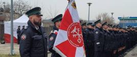 Funkcjonariusze Urzędu Celno-Skarbowego stoją ze sztandarem obok stoją w rzędzie pozostali funkcjonariusze.