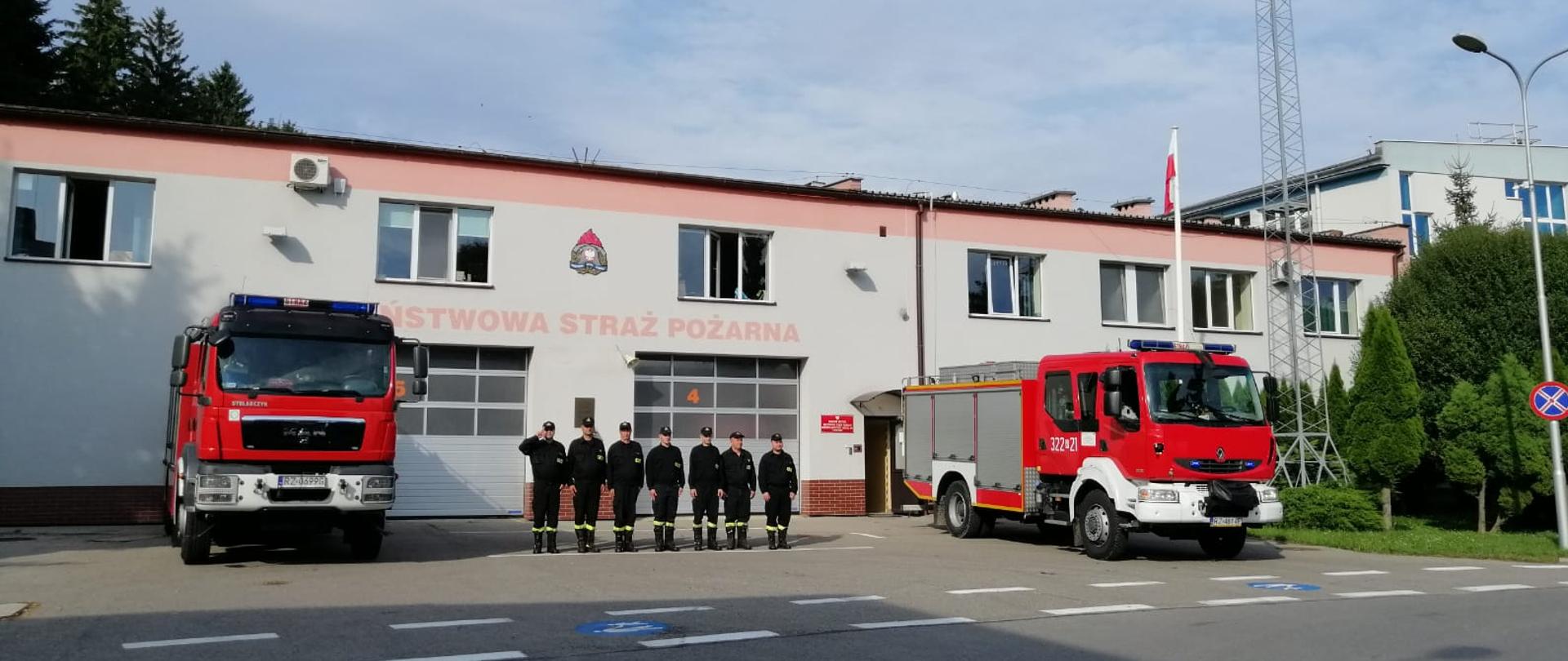 Na zdjęciu widoczna na zbiórce zmiana służbowa w ubraniu koszarowym - 7 strażaków, dowódca oddaje honor. Po ich obydwu stronach stoją dwa samochody pożarnicze z włączonymi światłami alarmowymi. W tle remiza strażacka.