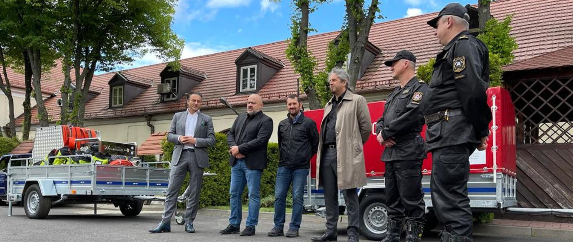 Na zdjęciu widać sześciu mężczyzn- dwóch z nich ubranych jest w mundury. Osobami na zdjęciu są przedstawiciele firmy Ghelamco Poland Sp. z o.o. oraz dwóch strażaków - Komendant Miejski PSP m.st. Warszawy st. bryg. Leszek Smuniewski oraz Dowódca JRG 4 bryg. Sławomir Sierpatowski. Wszystkie osoby na zdjęciu stoją na tle dwóch przyczepek, na których znajdują się zestawy pompowe służące do podawania wody na wysokości 200m+.