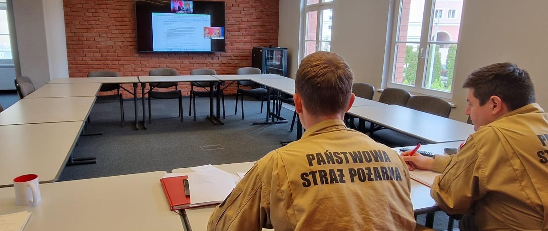 dwóch strażaków siedzi przy stole na przeciwko telewizora 