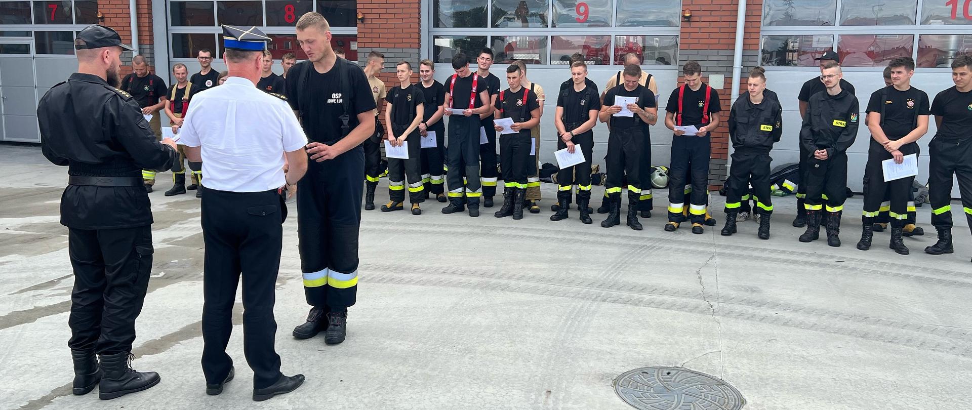 Na zdjęciu widać strażaków ustawionych w dwuszeregu na tle budynku strażnicy, przed frontem pododdziału wiać funkcjonariuszy PSP wręczających zaświadczenia ukończenia szkolenia