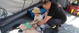 Zdjęcie wykonane na Placu Jagiellońskim w Radomiu podczas rodzinnego pikniku zdrowia zorganizowanego przed urzędem miejskim w Radomiu. Na zdjęciu widać wnętrze namiotu. W namiocie znajduje się strażak oraz dziecko. Na ziemi leży manekin – fantom do nauki pierwszej pomocy przedmedycznej. Strażak uczy dziecko jak udzielać pomocy osobom jej potrzebującym. Obok nich po prawej stronie widać czerwoną torbę w której znajdują się różnego rodzaju opatrunki, bandaże, kołnierze i tym podobne rzeczy przeznaczone do udzielania pierwszej pomocy.