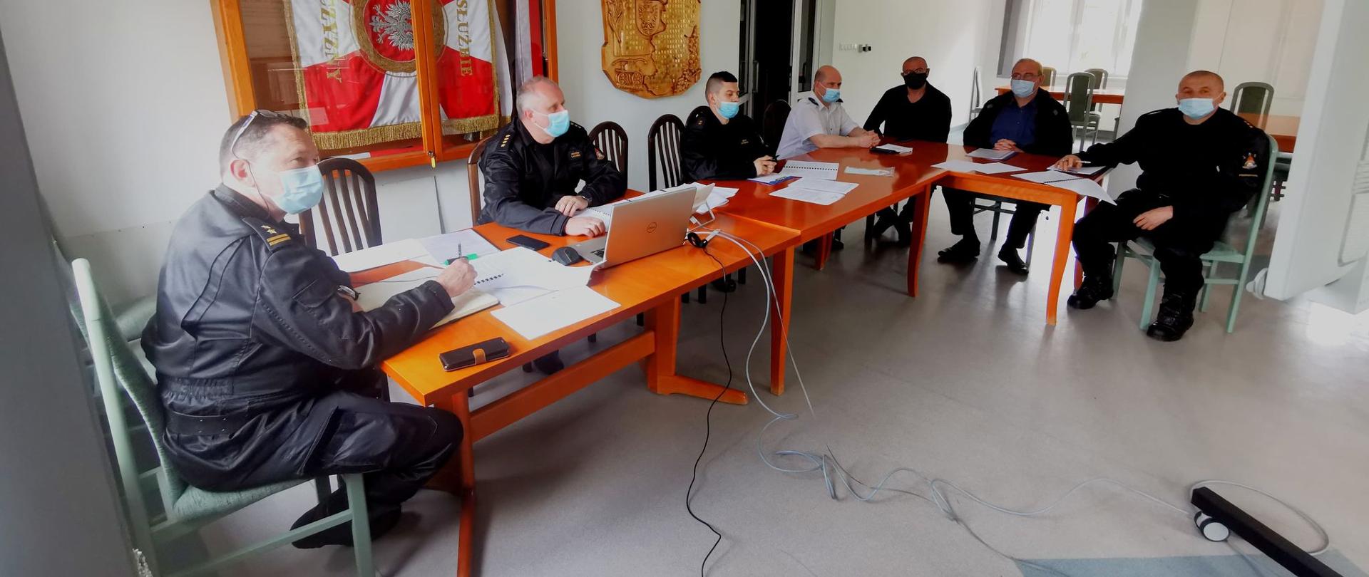 Zdjęcie przedstawia czterech funkcjonariuszy Państwowej Straży Pożarnej w Przeworsku oraz trzech druhów.
Uczestnicy wideokonferencji znajdują się w jasnym pomieszczeniu, na ścianie za strażakami widnieje gablota ze sztandarem.
Wszyscy siedzą przy stole w kształcie litery L, a twarze mają zasłonięte maseczką.
Na stole ułożone są dokumenty oraz komputer.