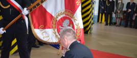 Zdjęcie przedstawia komendanta klękającego na prawe kolano i całującego sztandar, na ziemi czerwony dywan, w tle goście