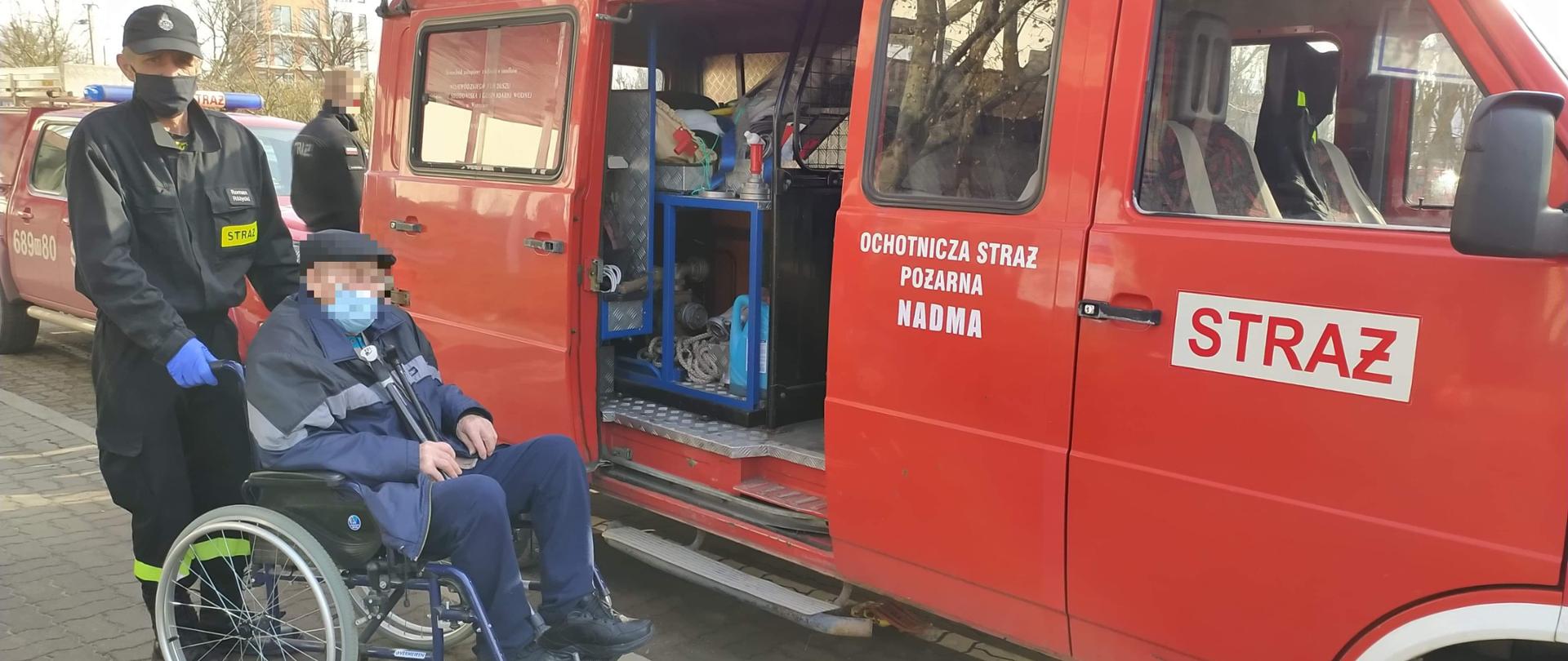 Na zdjęciu starszy mężczyzna na wózku inwalidzkim przed samochodem Ochotniczej Straży Pożarnej. Samochód typu bus czeka z otwartymi drzwiami. Wózek przytrzymywany jest przez strażaka w mundurze.