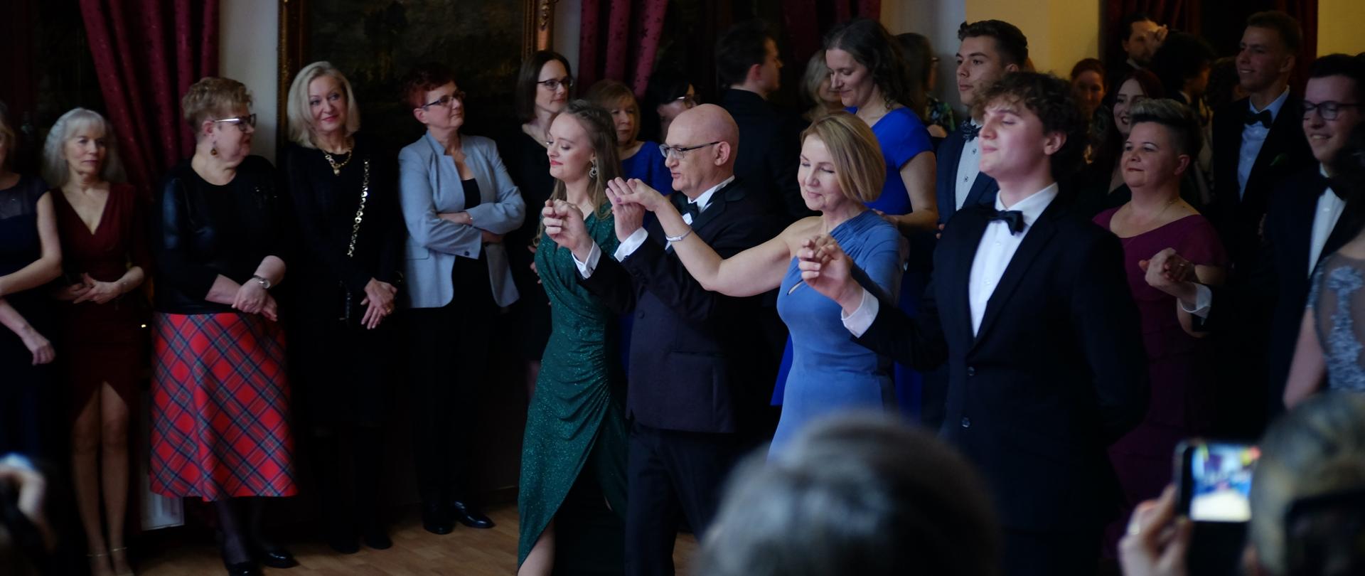 zdjęcie przedstawia dyrekcję szkoły, wychowawczynię klasy oraz uczniów i uczennice klasy maturalnej podczas innej figury Poloneza na sali bankietowe,j w tle inni nauczyciele obserwujący taniec