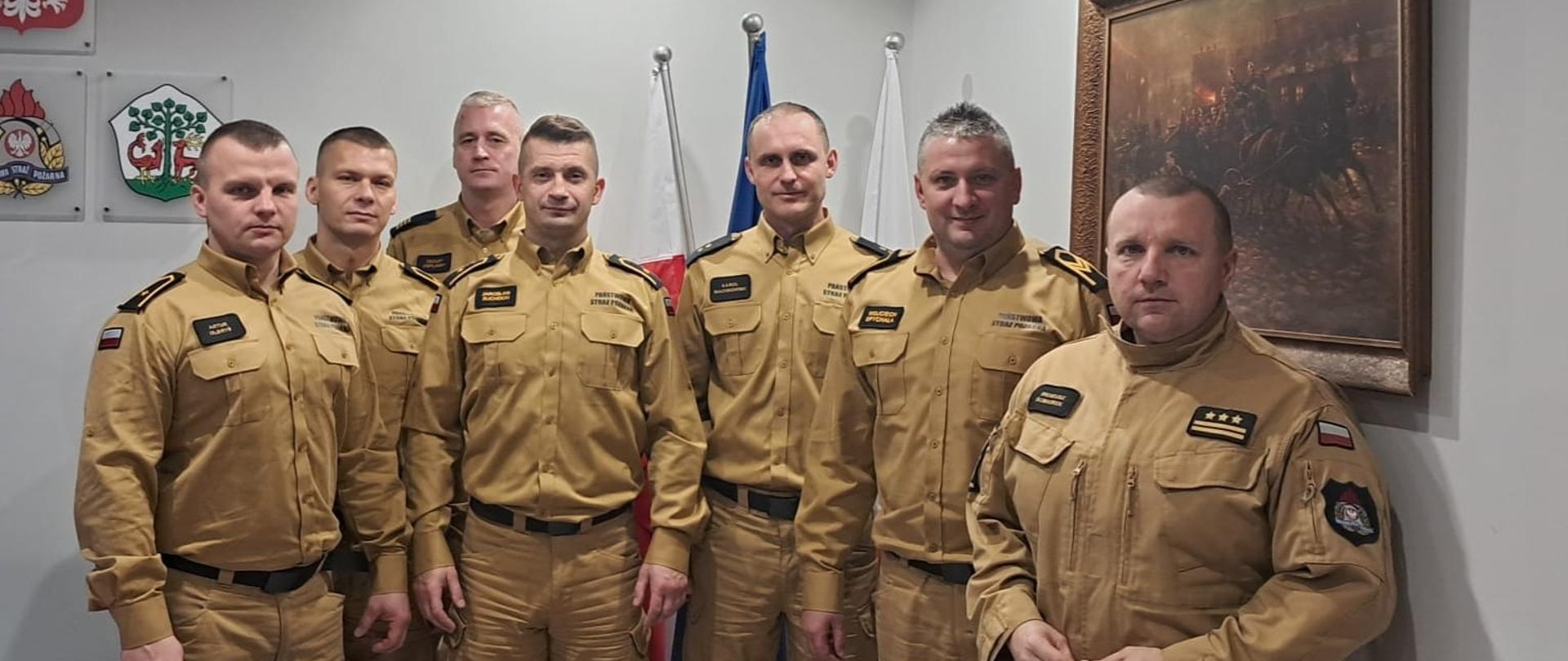 Na zdjęciu w gabinecie komendanta siedmiu strażaków w jasnych mundurach.