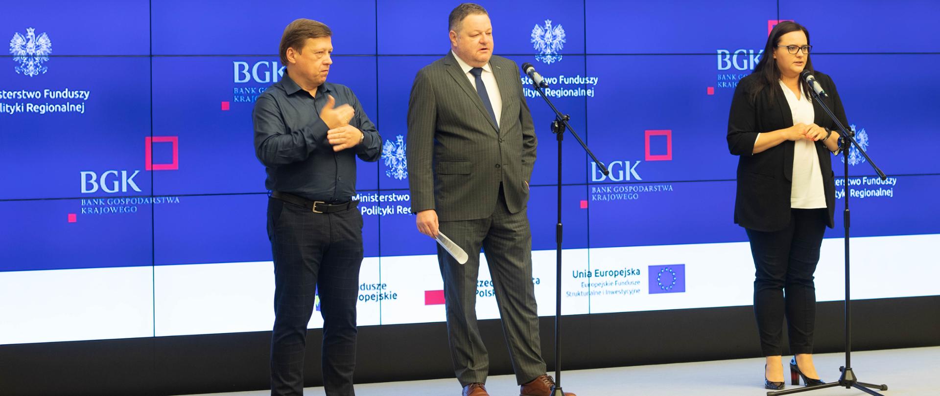Na sali konferencyjnej przed mikrofonami od prawej stoją minister M. Jarosińska-Jedynak i Przemysław Cieszyński, członek zarządu BGK. Obok tłumacz migowy. Za nimi granatowe tło z napisem Dodatkowe 1,1 mld zł z Funduszy Europejskich na pożyczki dla przedsiębiorców.