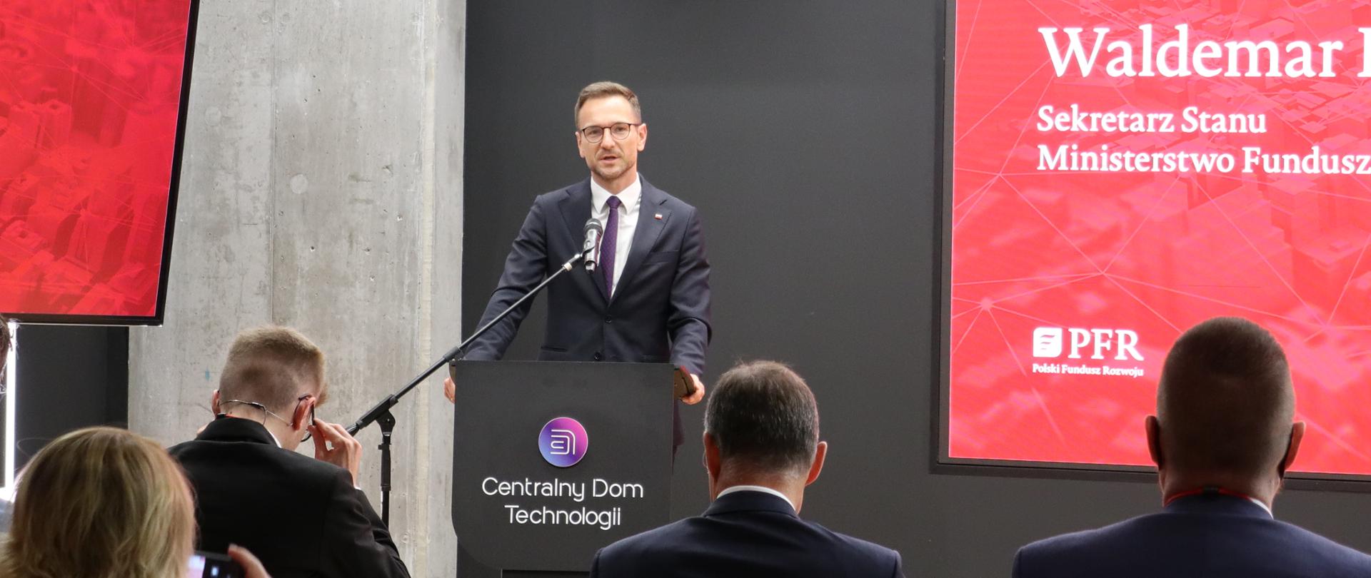 Wiceminister Waldemar Buda stoi w mównicy. Przed nim na krzesłach siedzą ludzie. Za nim ekran z lopgotypem PFR.