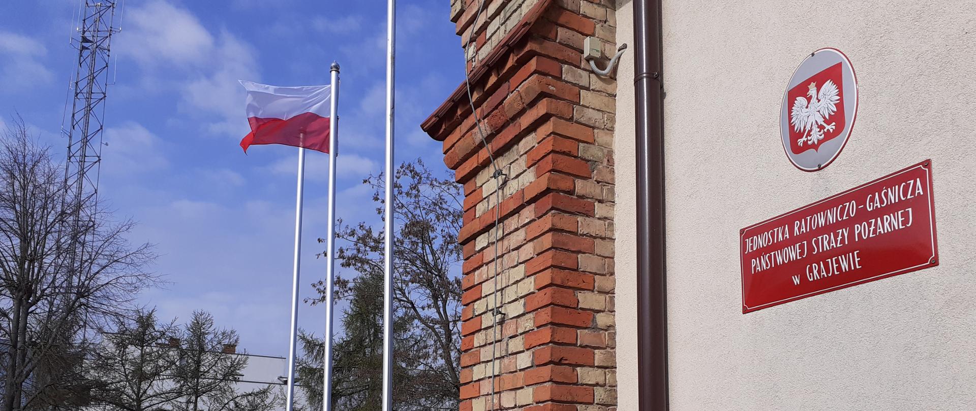 Polska flaga powiewa na maszcie/ pod masztem stoi poczet flagowy. Z prawej ściana budynku a na niej godło i tabliczka informacyjna.